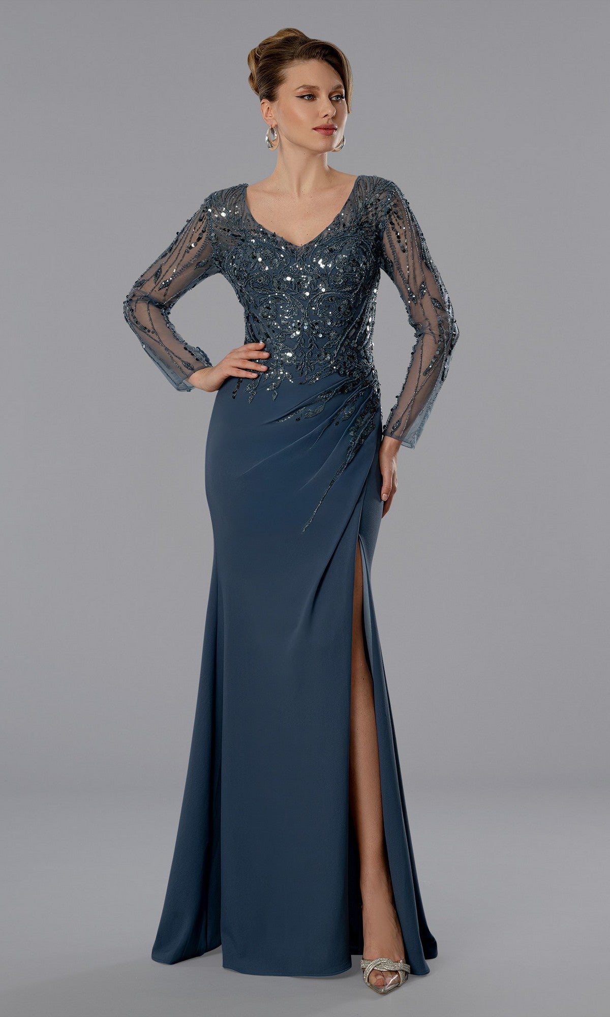 A woman in the Stella Couture 25371 long formal dress in charcoal, with a V-neck, sequin-embellished sheer long sleeves, side slit, and silver heels, stands against a plain gray background.